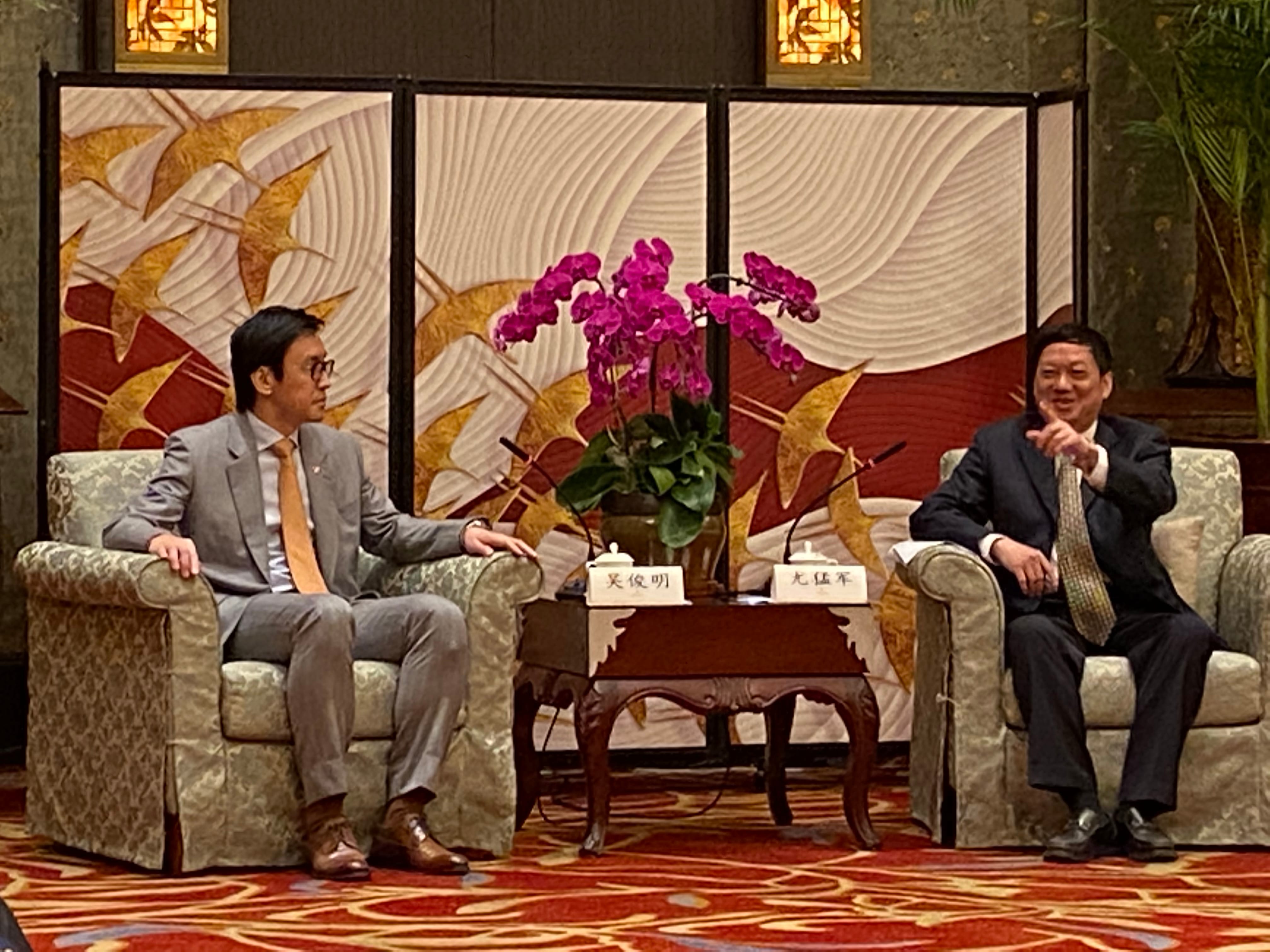 Two men in suits seated in armchairs, with a table and floral arrangement between them.
